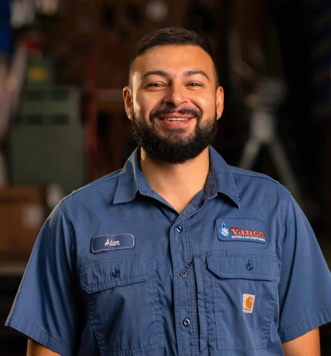 A Man With A Beard Is Smiling And Wearing A Blue Work Shirt With A Valco Mechanical Logo, Standing In A Workshop.