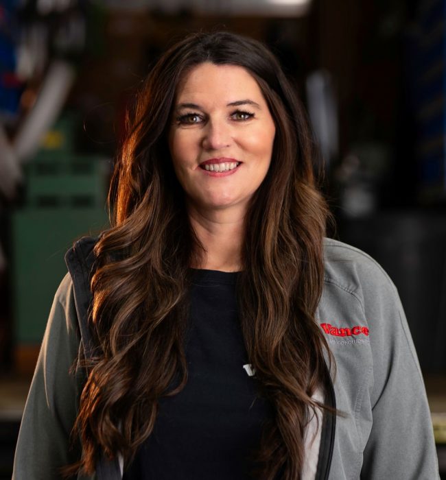 A Woman With Long Brown Hair Wearing A Gray Jacket Stands Indoors, Facing The Camera, With Workshop Equipment Visible In The Blurred Background—Perfect For An &Quot;About Us&Quot; Introduction.