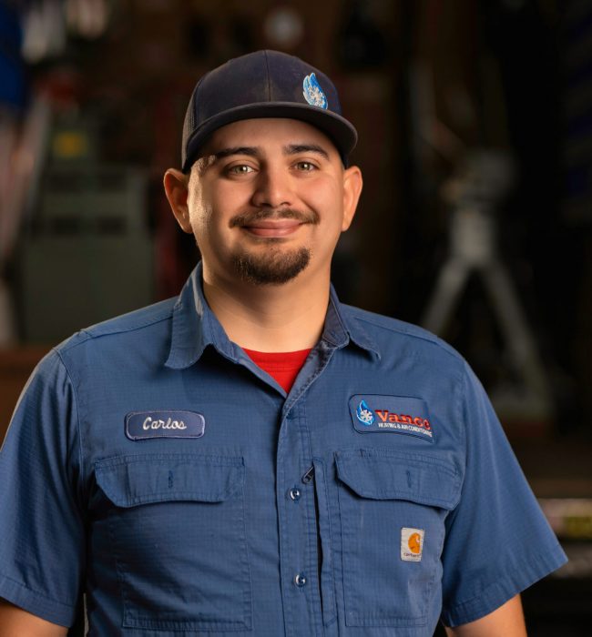 A man wearing a blue work shirt and cap stands indoors, facing the camera and smiling. His shirt displays a patch with the name “Carlos,” another with a company logo, making him a friendly face you'll meet on our About Us page.