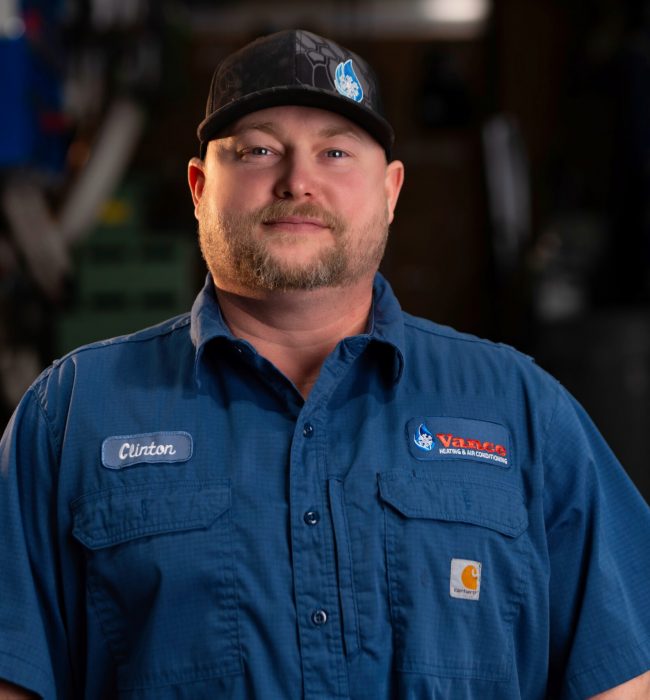 A man in a blue work shirt and black cap stands indoors, facing the camera. His shirt has a name tag labeled "Clinton." The background is blurred.