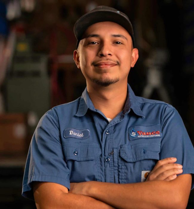 A Man In A Blue Work Shirt And Cap Stands With Arms Crossed, Smiling At The Camera In What Appears To Be A Workshop Or Garage Setting—Perfectly Capturing The Spirit Of Our Team On The About Us Page.
