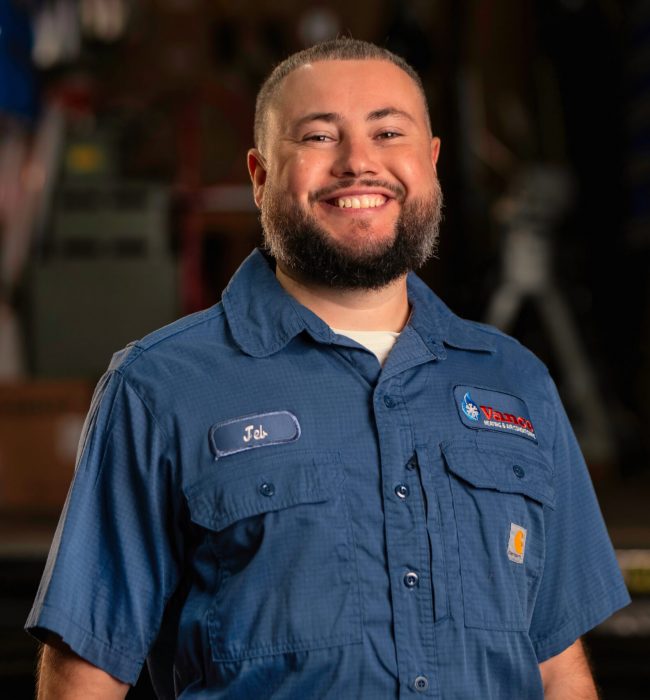 A man wearing a blue work shirt with a name tag that says "Job" smiles while standing indoors, with industrial equipment in the background, embodying the spirit of our about us story.
