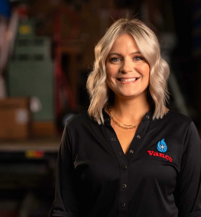 A Woman With Blonde Hair Wearing A Black Vance Company Shirt Stands Smiling In Front Of A Blurred Industrial Background, Representing The Friendly Faces Behind Our About Us Story.