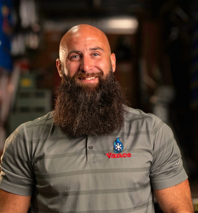A man with a bald head and long beard, wearing a gray striped polo shirt with "Vance" embroidered on it, sits smiling in an indoor industrial setting—capturing the welcoming spirit of our about us story.